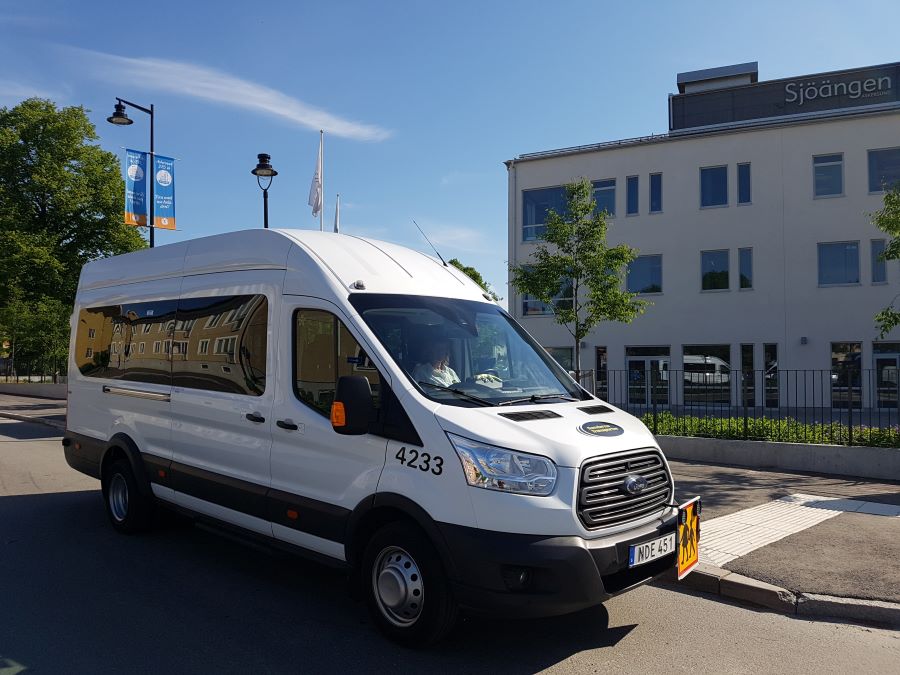 Vit minibuss som kör skolskjuts vid resecentrum med kulturhuset Sjöängen i Askersund i bakgrunden