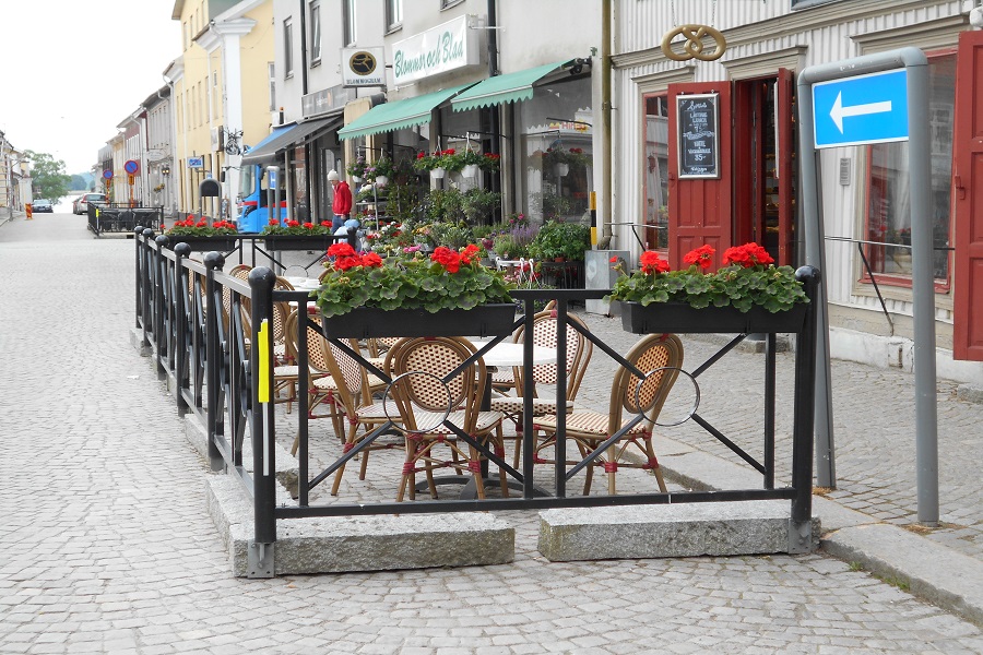 Uteservering med stolar och bord på en gata i anslutning till café. Blomlådor är uppsatta på räckena.