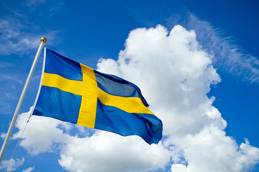 Svensk flagga hissad i flaggstång med blå himmel och vita moln som bakgrund.