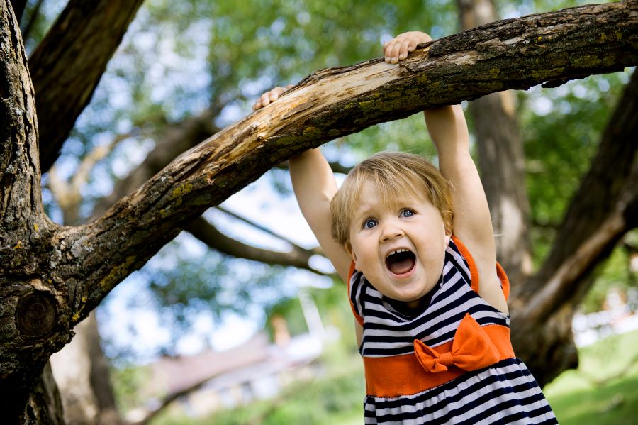Ett litet barn hänger i händerna i ett klätterträd. Barnet är cirka två år, har en svart/vitrandig klänning och det verkar som att det är både roligt och väldigt spännande att hänga i trädet.
