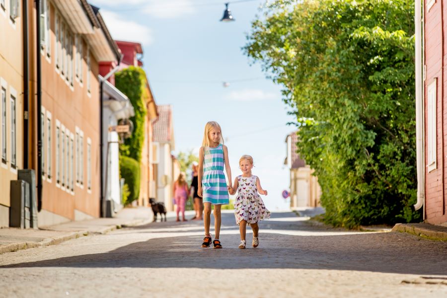 Två små flickor i sommarklänningar går hand i hand på gatstensgata mellan hus i centrala Askersund. Det är soligt och ser ut att vara en fin och varm sommardag.