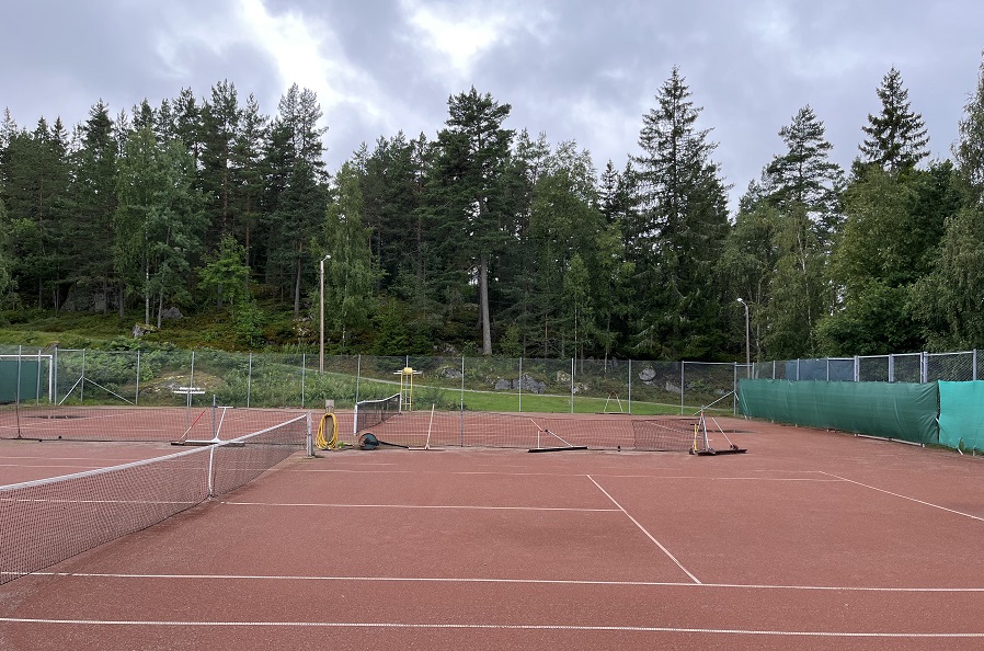 Två tennisbanor med nät på grusunderlag, ett staket inhägnar banorna. Gröna träd i bakgrunden.